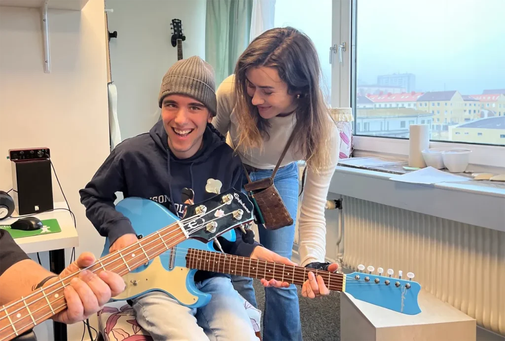 Deltagare och handledare spelar gitarr tillsammans. Deltagare kollar in i kameran med ett stort leende.